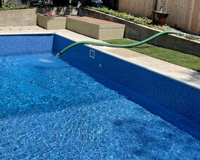 Brand new blue vinyl pool liner being filled with water beside a paver deck and a cedar fence